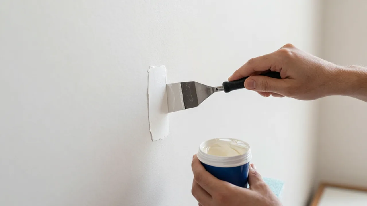 Manos aplicando compuesto blanco sobre una pared con una pequeña espátula metálica.