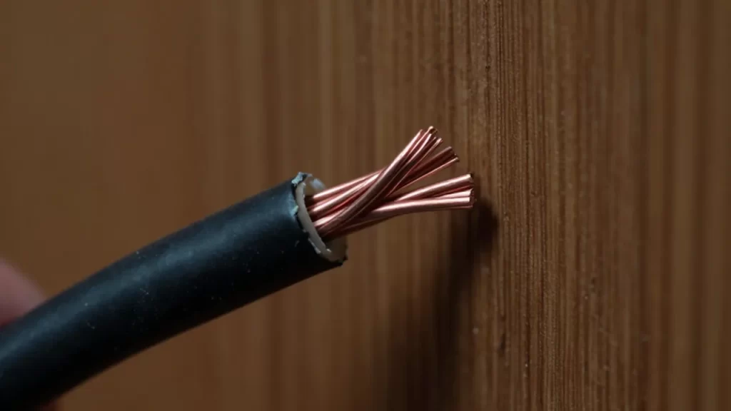 Cable eléctrico con aislamiento negro deshilachado y filamentos de cobre expuestos detrás de un panel de madera.