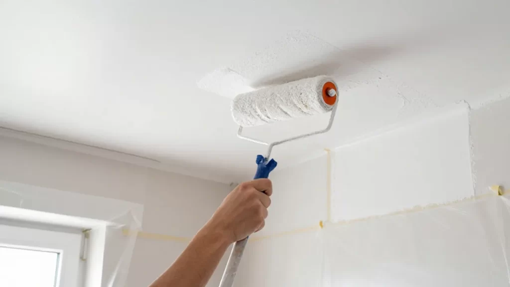 Persona utilizando un rodillo para pintar de blanco un techo con superficies protegidas.