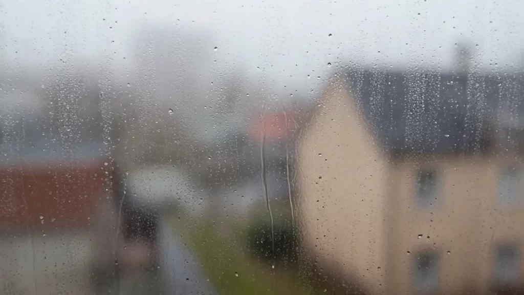 Gotas de condensación sobre el cristal de una ventana con un interior acogedor desenfocado al fondo.