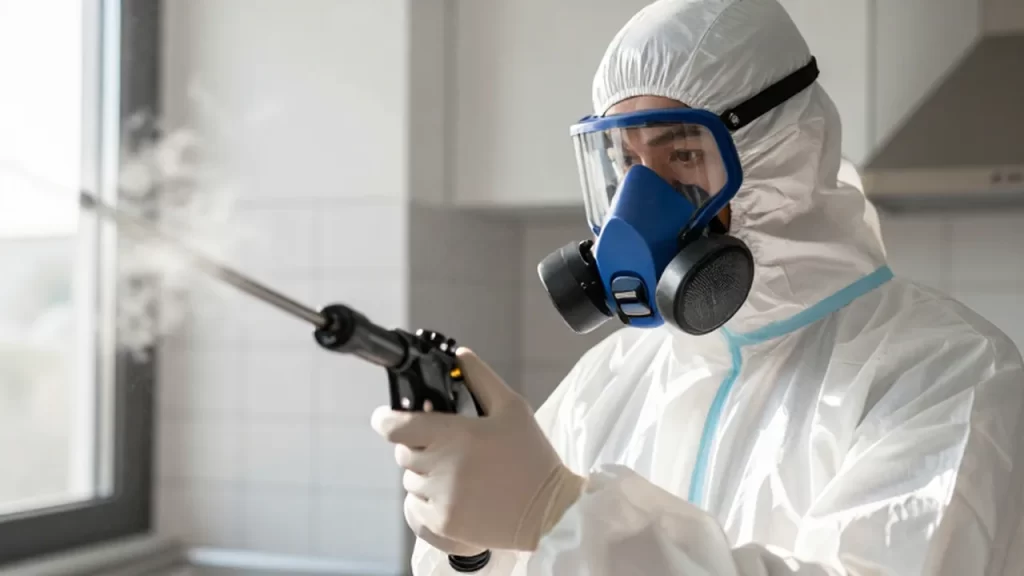 Técnico de control de plagas con traje protector y mascarilla usando un pulverizador en una cocina.